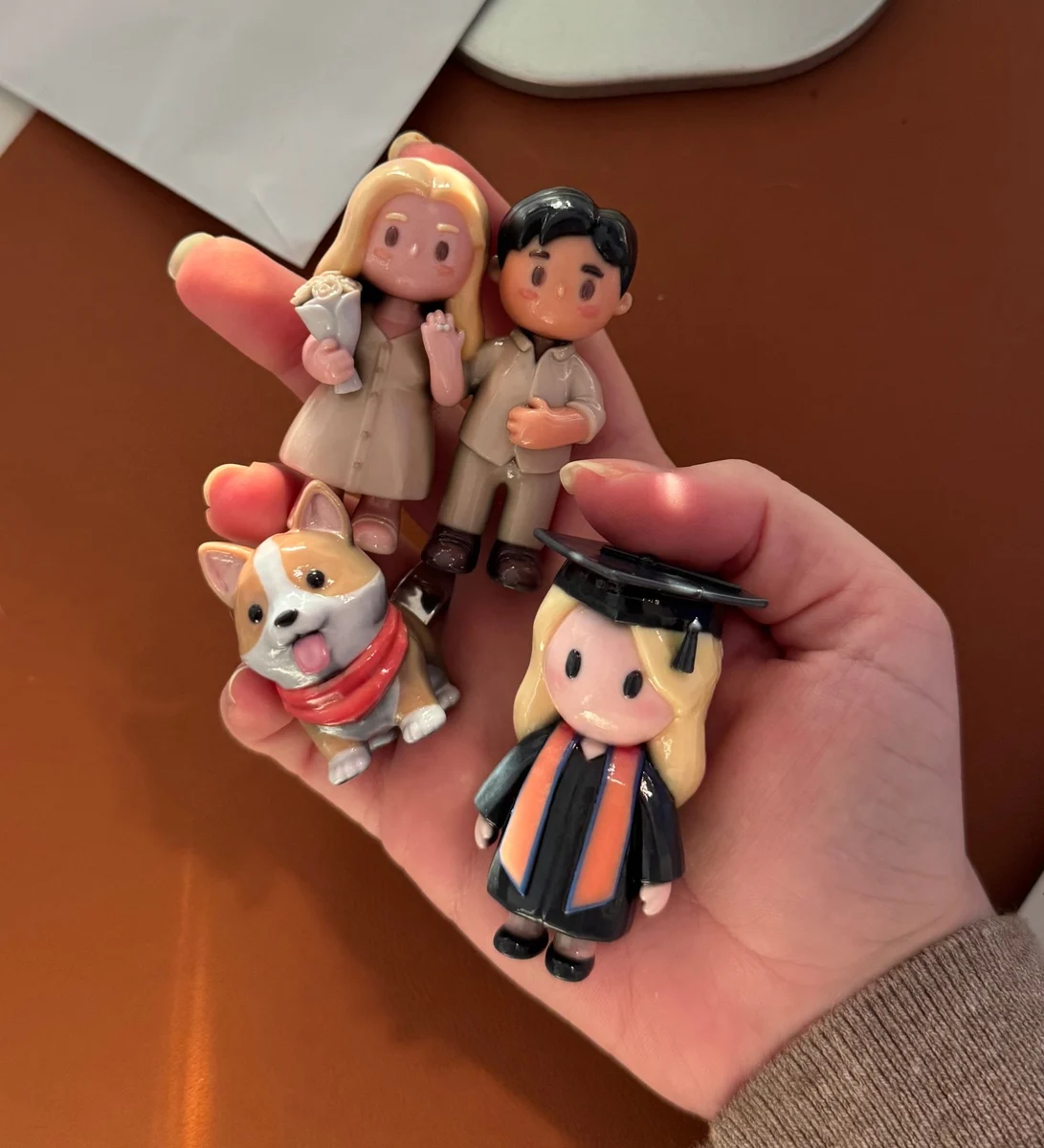 A hand holding custom 3D-printed figurines of a couple, a corgi and a graduate